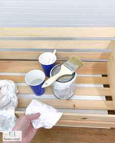 a person holding a paint brush next to two cups on a wooden shelf with white paper