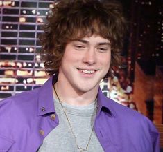 a young man with curly hair wearing a purple jacket and smiling at the camera while standing in front of a tv screen