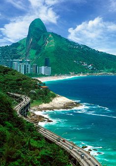 an aerial view of the ocean, mountains and road with cars driving on it in front of a large mountain