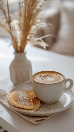 a cup of cappuccino and some pancakes on a plate with a vase in the background