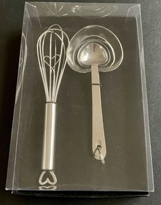 two kitchen utensils in a clear box on a black counter top, one has a whisk and the other is an egg beater