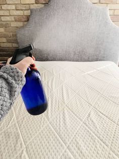 a person holding a blue bottle on top of a white bed with a brick headboard