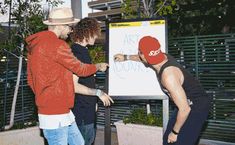 three men standing around a white board with writing on it and one man wearing a red hat