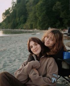 two women sitting on a beach next to each other with trees in the back ground