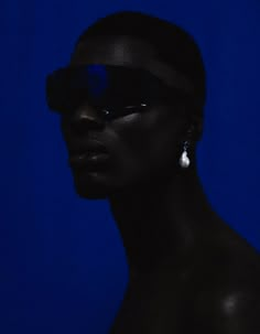 a woman with sunglasses on her head and dark skin wearing white earrings, against a blue background