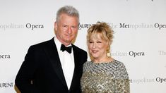 a man and woman standing next to each other in front of a white wall with the metropolitan opera logo on it