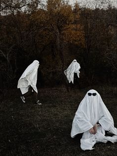 two ghostly people in white cloths are sitting on the ground with trees behind them