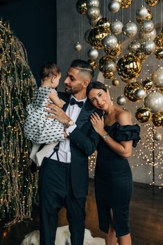 a man in a tuxedo holding a baby and standing next to a christmas tree