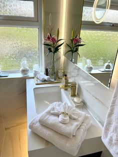 towels are stacked on the counter in front of a bathroom mirror and sink with two large windows