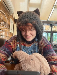 a woman in overalls holding a teddy bear wearing a knitted hat with ears