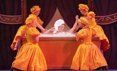 five women in yellow dresses and hats stand around a bathtub with foam on it