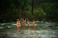 four women in bathing suits standing on the edge of a river with their arms up