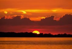 the sun is setting over water with clouds in the sky and trees on the shore