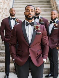 A group of groomsmen standing on stone steps, each wearing matching burgundy tuxedo jackets with black satin shawl lapels, black dress pants, white shirts, and black bow ties. Each man has a boutonnière pinned to his jacket, giving a cohesive and formal look to the group. Red Velvet Suit Mens Wedding, Black And Burgundy Groomsmen, Maroon Groomsmen Attire, Maroon Groom Suit, Maroon Grooms Suit, Burgundy Suit Men, Red Velvet Suit, Red Suits, Sophisticated Men