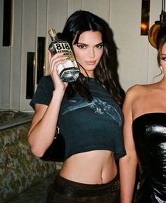 two beautiful women standing next to each other in front of a couch and holding bottles