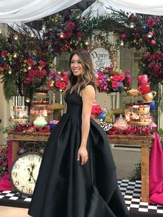 a woman wearing a black dress standing in front of a table with cakes and flowers