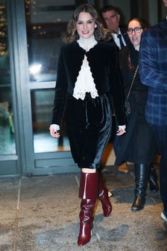 a woman in black dress and boots walking down the street with other people behind her