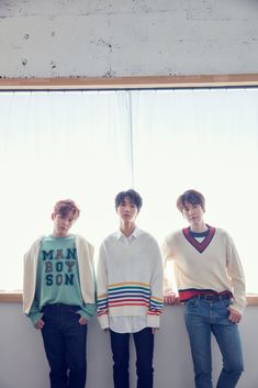 three young men standing next to each other in front of a window, wearing sweaters and jeans