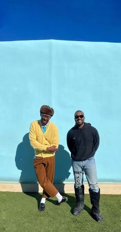 two men standing next to each other in front of a blue wall and grass area