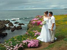 a bride and groom standing on the edge of a cliff overlooking the ocean with wildflowers