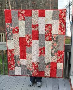 a person holding up a red and gray quilt on a porch with trees in the background