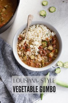 a white bowl filled with vegetable gumbo next to a pot of rice and spoon