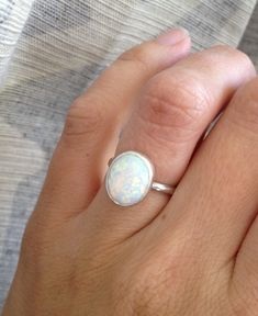 An oval simulated opal has been set in sterling silver with a handmade ring band from sterling silver half dome wire. This ring is simple, elegant and luminescent. The opal changes colors when it catches the light showing pinks, blues, oranges, and whites. The opal is 12mm x 10mm in size and is a perfect addition to any collection. Made to order. Available in both a bright sterling silver finish or a dark patina finish that gives the ring a little more edge. Opal is the birthstone for October! A Adjustable Oval Opal Ring Minimalist Style, Adjustable Oval Opal Jewelry, Minimalist Silver Opal Ring, White Opal Oval Cabochon Ring, Minimalist Adjustable Oval Opal Ring, Sterling Silver Oval Opal Cabochon Ring, Minimalist Oval Opal Ring In Sterling Silver, Minimalist Sterling Silver Oval Opal Ring, Minimalist Sterling Silver Opal Ring Oval