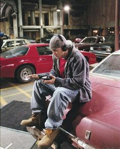 a man sitting on the hood of a red car looking at his cell phone while listening to headphones