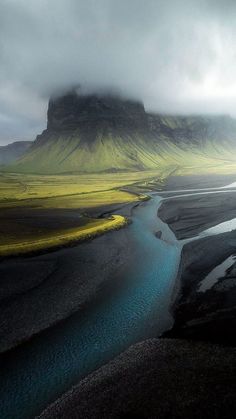 a river running through a lush green field next to a mountain covered in fog and clouds