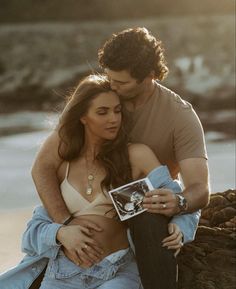 a man holding a woman on the beach while she is leaning against her chest and looking at an album