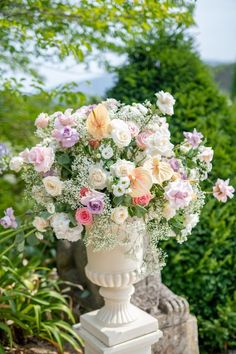 a white vase filled with lots of flowers on top of a stone wall next to bushes