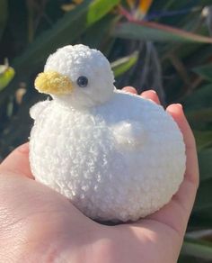 a small white stuffed animal sitting on top of someone's hand in front of some plants