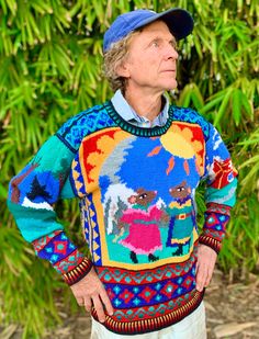 an old man wearing a colorful sweater and hat standing in front of some bushes with his hands on his hips