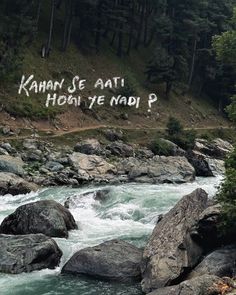 graffiti written on the side of a mountain above a river with rocks and trees in the foreground