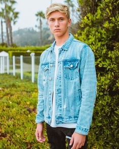 a young man wearing a jean jacket and black pants standing in front of bushes with his hands in his pockets