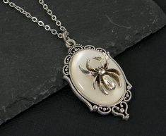 Add a bit of goth mystique with this silver spider necklace. This white and silver spider necklace has a victorian style with a pendant that features a three dimensional spider over a white oval. An ornate silver filigree frame holds the white oval. The perfect gift any man or women that represents a strong and powerful personality. Shop now and show off your gothic style! Pendant is 1 5/8 inches tall and 7/8 inches wide. Silver plated necklace chain - 18, 24 or 30 inches long. Also available in White Gothic Necklace Gift, Spider Clothes, Powerful Personality, Goth Spider, Spider Pendant, Filigree Frame, White Goth, Jewelry 2023, Vintage Spider