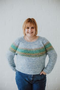 a woman standing in front of a white brick wall wearing a blue sweater and jeans