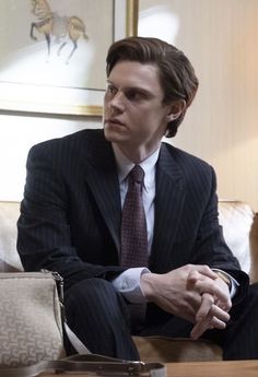 a man in a suit and tie sitting on a couch with his hands folded out