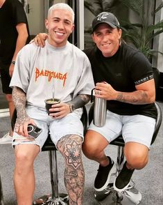 two men sitting next to each other with tattoos on their arms and legs, one holding a drink in his hand