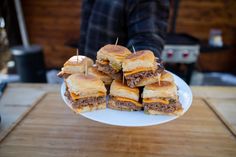 a white plate topped with mini sandwiches on top of a wooden table next to a person