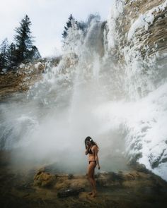 a woman standing in front of a waterfall with steam coming out of the water and trees around her