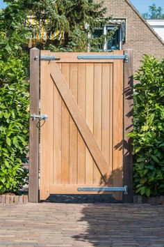 an open wooden gate in front of a brick walkway and shrubbery on the side of a house