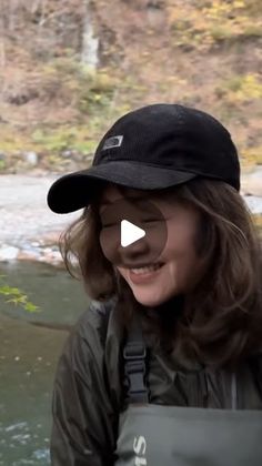 a woman wearing a black hat standing in front of a river and smiling at the camera