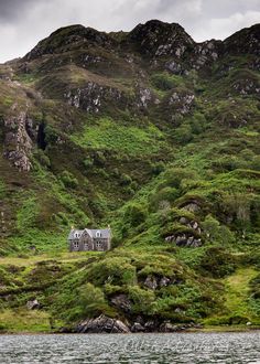 an old house on the side of a mountain with green grass and bushes around it