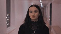 a woman with long black hair standing in an empty hallway, looking at the camera