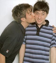 two young men are kissing each other in front of a white wall and one is wearing a striped shirt