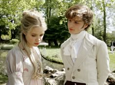 two young people standing next to each other in front of a pond and trees with green leaves