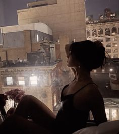 a woman sitting on top of a bed looking out the window at buildings in the distance