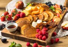 french toast with fruit and syrup on a wooden cutting board next to fresh berries, raspberries, and apples