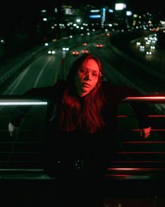 a woman standing on a bridge at night with her arms over her shoulder and looking off into the distance